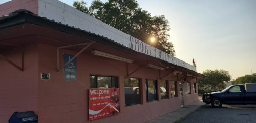 Exterior of a pink building with a sign reading "Smokey's" and a "Welcome" banner, parked vehicles nearby.